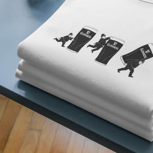 May include: White t-shirt featuring a black graphic of three figures carrying large Guinness pints. The Guinness logo is visible on each pint. The shirt is folded and stacked on a blue surface.