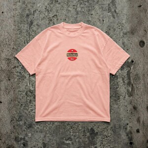 Pu&ograve; includere: Una maglietta rosa chiaro con girocollo e maniche corte. La maglietta presenta un logo circolare con una stella rossa, la parola "Heineken" e la parola "bier" in un design rosso e oro.