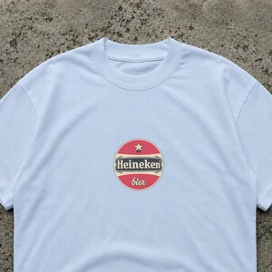 Puede incluir: Una camiseta blanca con un logotipo circular que dice "Heineken bier". El logotipo es rojo, blanco y beige, con una estrella en la parte superior. La camiseta tiene cuello redondo y mangas cortas.