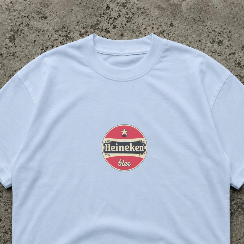 Puede incluir: Una camiseta blanca con un logotipo circular que dice "Heineken bier". El logotipo es rojo, blanco y beige, con una estrella en la parte superior. La camiseta tiene cuello redondo y mangas cortas.