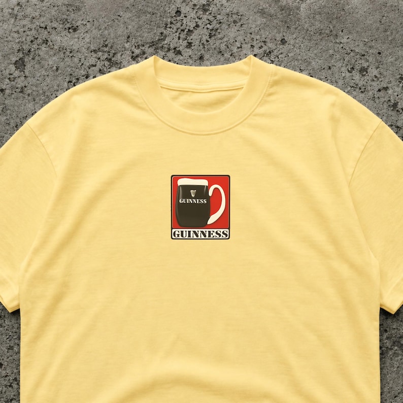 May include: A light yellow t-shirt featuring a graphic of a pint of Guinness stout. The graphic is in a red square with a white handle and the word "Guinness" in a white font. The shirt is made of a soft material.