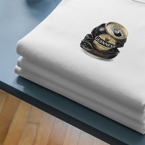 May include: A stack of three folded white shirts on a blue surface. A crushed black and gold Guinness beer can rests on top of the shirts. The can's label is visible, with the words "Guinness Original".
