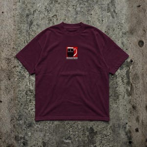May include: Burgundy t-shirt featuring a small, square graphic of a Guinness pint with the brand name below. The graphic is in black, white, and red. The shirt has a crew neck and short sleeves.
