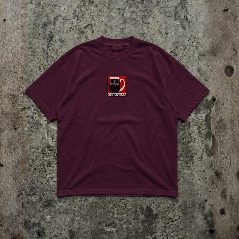 May include: Burgundy t-shirt featuring a small, square graphic of a Guinness pint with the brand name below. The graphic is in black, white, and red. The shirt has a crew neck and short sleeves.