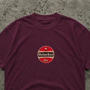 Pode incluir: T-shirt bord&ocirc; com um logotipo circular. O logotipo diz "Heineken bier" em texto branco sobre um fundo vermelho, com uma estrela dourada. A t-shirt &eacute; de cor lisa.