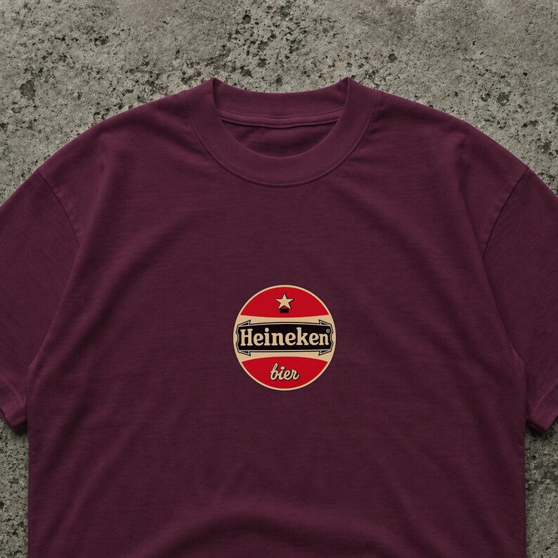 Pode incluir: T-shirt bord&ocirc; com um logotipo circular. O logotipo diz "Heineken bier" em texto branco sobre um fundo vermelho, com uma estrela dourada. A t-shirt &eacute; de cor lisa.
