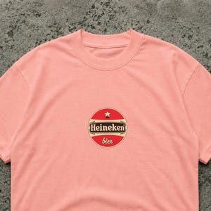 May include: A light pink t-shirt featuring a circular logo with the text "Heineken" in white on a brown background, with a red border and a star at the top. The word "bier" is written in red below the logo.