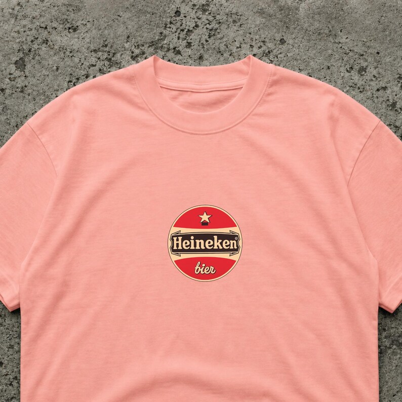 May include: A light pink t-shirt featuring a circular logo with the text "Heineken" in white on a brown background, with a red border and a star at the top. The word "bier" is written in red below the logo.