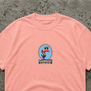 May include: A light pink t-shirt featuring a Guinness logo with a toucan holding two pints of stout. The logo is in a blue circle with the text "Guinness is good for you" below it.