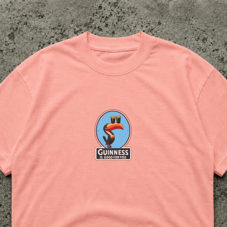 May include: A light pink t-shirt featuring a Guinness logo with a toucan holding two pints of stout. The logo is in a blue circle with the text "Guinness is good for you" below it.