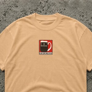 May include: A tan crew neck t-shirt featuring a small, rectangular graphic of a Guinness pint. The graphic is a black pint of stout with a white head, set against a red background, with the Guinness logo.