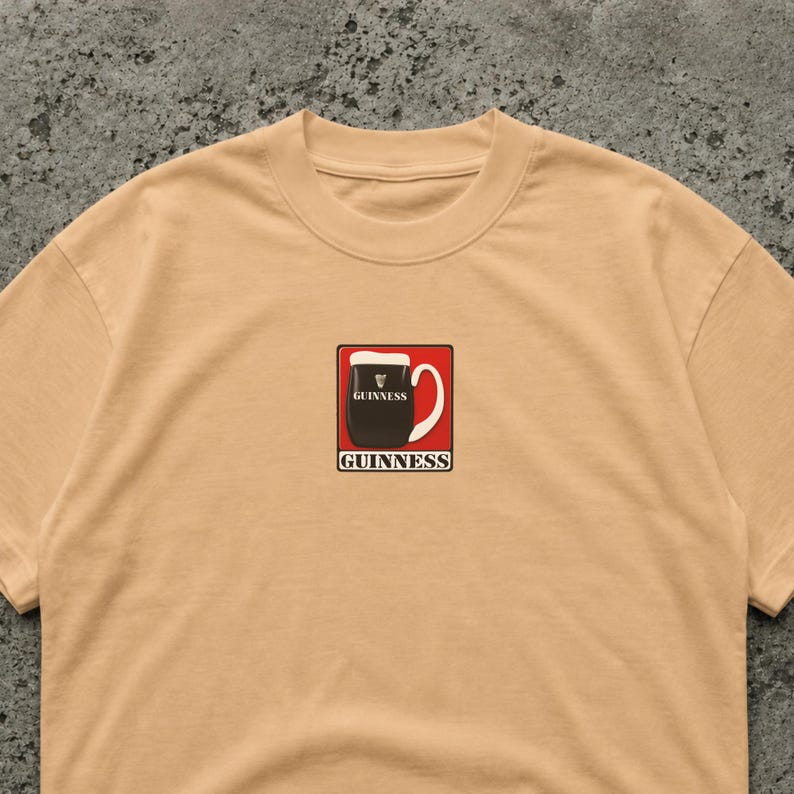 May include: A tan crew neck t-shirt featuring a small, rectangular graphic of a Guinness pint. The graphic is a black pint of stout with a white head, set against a red background, with the Guinness logo.