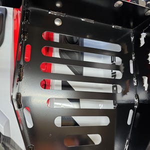 Puede incluir: Una cesta de almacenamiento de metal negro con ranuras horizontales y esquinas redondeadas. La cesta est&aacute; montada en un veh&iacute;culo, con detalles en rojo y blanco visibles en el fondo. Soldaduras y remaches aseguran la estructura.