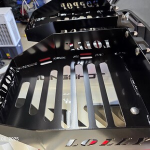 Puede incluir: Cestas de almacenamiento de metal negro con letras recortadas. Las cestas tienen forma rectangular con ranuras verticales y est&aacute;n montadas en un veh&iacute;culo. Los textos "TONKA" y "BOBBO" son visibles. El dise&ntilde;o es industrial.