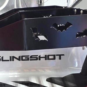 Puede incluir: Accesorio de metal negro con recortes de murci&eacute;lagos, montado sobre una superficie blanca con la palabra "SLINGSHOT" en negro. El accesorio est&aacute; fijado a una estructura de soporte negra, probablemente para un veh&iacute;culo.