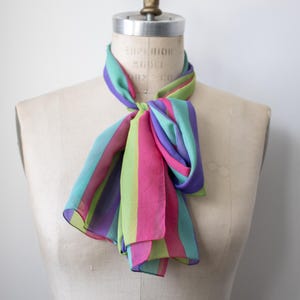 May include: A lightweight scarf featuring vertical stripes in vibrant shades of magenta, lime green, turquoise, and purple. The scarf is tied in a simple knot around the neck of a beige dress form, displaying a semi-sheer fabric texture.