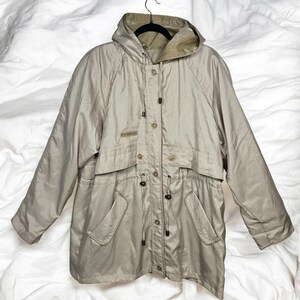 May include: A beige hooded jacket with a drawstring waist and multiple pockets. The jacket has a button-up front and a light-colored lining in the hood. The jacket is hanging on a black hanger against a white background.