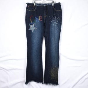 May include: Dark wash denim jeans with a flared leg. The jeans feature the word "LOVE" spelled out in colorful rhinestones, a rhinestone star, and a rhinestone design on the right leg. The jeans have a frayed hem and are hanging on a hanger.