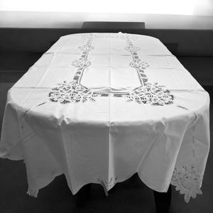 May include: A white oval tablecloth with intricate lace detailing. The lace design features floral and geometric patterns along the center and edges. The tablecloth is draped over a dark table, creating a contrast.