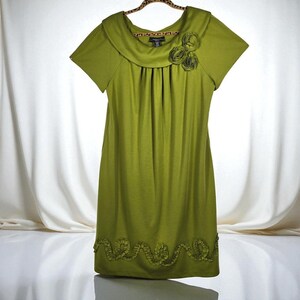 May include: A short-sleeved, olive green dress with a round neckline and a decorative floral embellishment on the chest. The dress features a gathered bodice and a ruffled hemline. The dress is hanging on a leopard print hanger.
