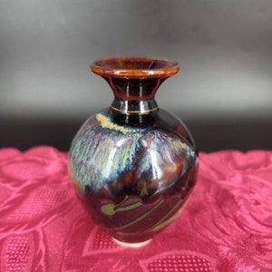 Op de afbeelding: Een kleine, ronde, donkerbruine keramische vaas met een brede, uitlopende opening. De vaas heeft een glanzende afwerking met iriserende blauwe, paarse en gouden accenten.