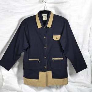 May include: Navy blue jacket with tan accents. The jacket features a tan collar, pocket details, and a tan hem. Gold-colored buttons and zipper pockets add detail to the design. The jacket is hanging on a metal hanger.