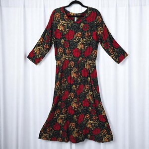 May include: A long-sleeved dress with a floral pattern in red, gold, and green on a black background. The dress has a V-neck and a gathered waist. The dress is hanging on a black hanger.