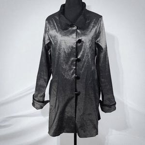 May include: A long, dark gray coat with a shiny, textured appearance. The coat features a classic collar, long sleeves with turned-up cuffs, and a row of four black buttons down the front. The coat is displayed on a black mannequin.