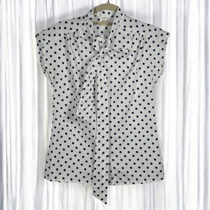 Peut inclure: Chemisier blanc à pois bleu marine. Le chemisier a des manches courtes et un détail de col noué. Le vêtement est suspendu à un cintre en bois sur fond blanc.