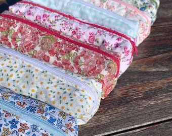 Puede incluir: Seis estuches de lápices con cremallera de colores con estampados florales. Los estuches están alineados sobre una superficie de madera.