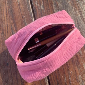 Op de afbeelding: Een roze, gewatteerde toilettas met een ritssluiting. De tas is open en gevuld met make-upkwasten en andere cosmetica.