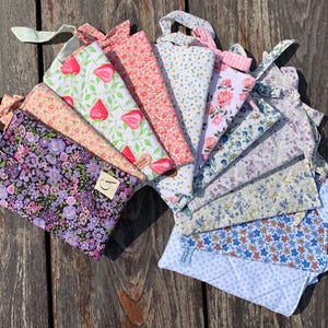 Könnte beinhalten: Eine Sammlung von Stoffbeuteln mit verschiedenen Blumen- und Fruchtmustern, darunter Erdbeeren und Rosen. Die Beutel sind fächerförmig angeordnet und zeigen eine Vielzahl von Farben und Designs. Jeder Beutel hat eine Schlaufe zum Aufhängen.