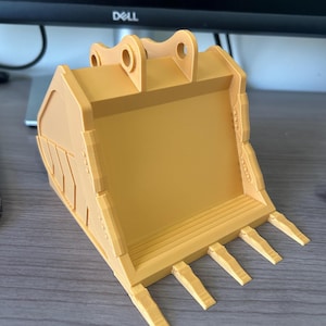 Puede incluir: Un cuchar&oacute;n de excavadora amarillo impreso en 3D, con forma de pala y dientes en la parte inferior. El cuchar&oacute;n tiene dos agujeros circulares en la parte superior para la fijaci&oacute;n y est&aacute; dise&ntilde;ado con crestas y bordes detallados.