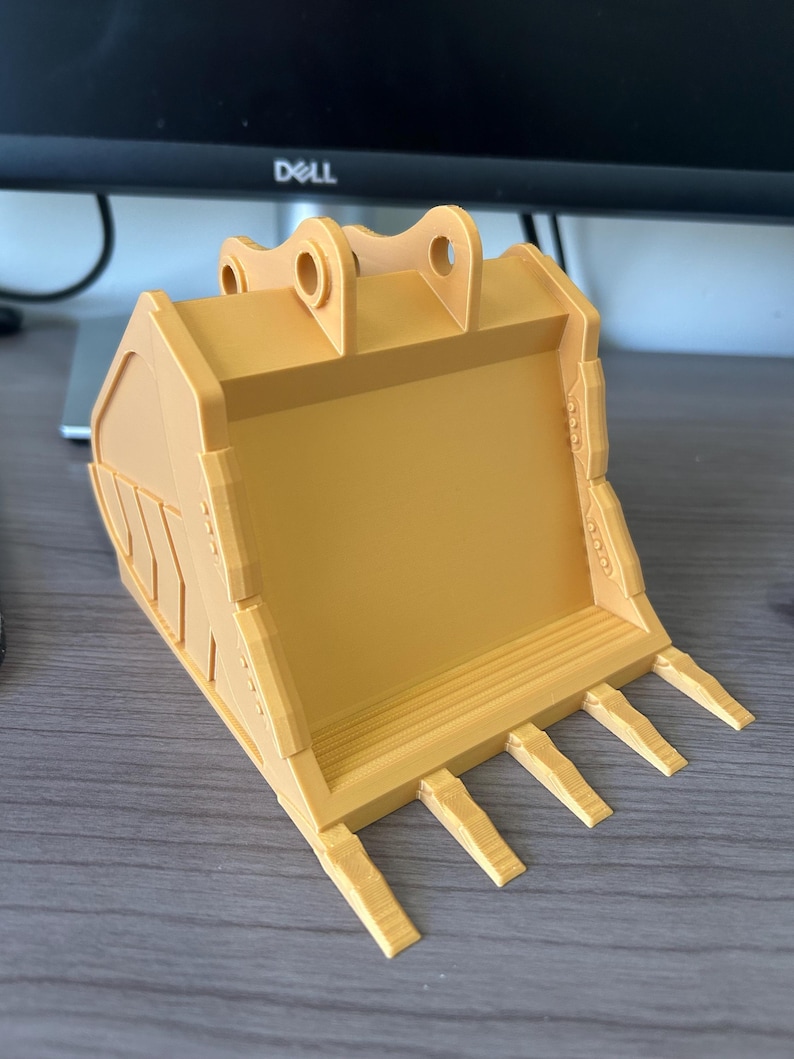 Puede incluir: Un cuchar&oacute;n de excavadora amarillo impreso en 3D, con forma de pala y dientes en la parte inferior. El cuchar&oacute;n tiene dos agujeros circulares en la parte superior para la fijaci&oacute;n y est&aacute; dise&ntilde;ado con crestas y bordes detallados.