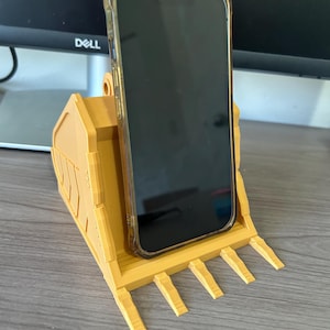 Puede incluir: Un soporte para tel&eacute;fono amarillo impreso en 3D con forma de cuchara de excavadora. Un tel&eacute;fono inteligente negro con una funda transparente descansa en el soporte. El soporte tiene una base plana con cinco proyecciones en forma de dientes.