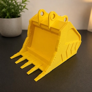 May include: A bright yellow excavator bucket model featuring a reinforced cutting edge with four distinct teeth, side wall ribbing, and two top mounting lugs. The construction equipment replica is rendered in a solid, matte yellow finish.