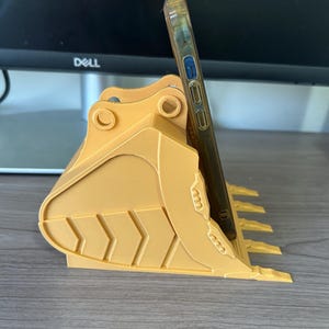 Puede incluir: Un soporte para tel&eacute;fono amarillo impreso en 3D con forma de cuchara de excavadora. Tiene una ranura para sujetar un tel&eacute;fono en posici&oacute;n vertical. El tel&eacute;fono est&aacute; en una funda transparente. El soporte est&aacute; sobre una superficie de madera.