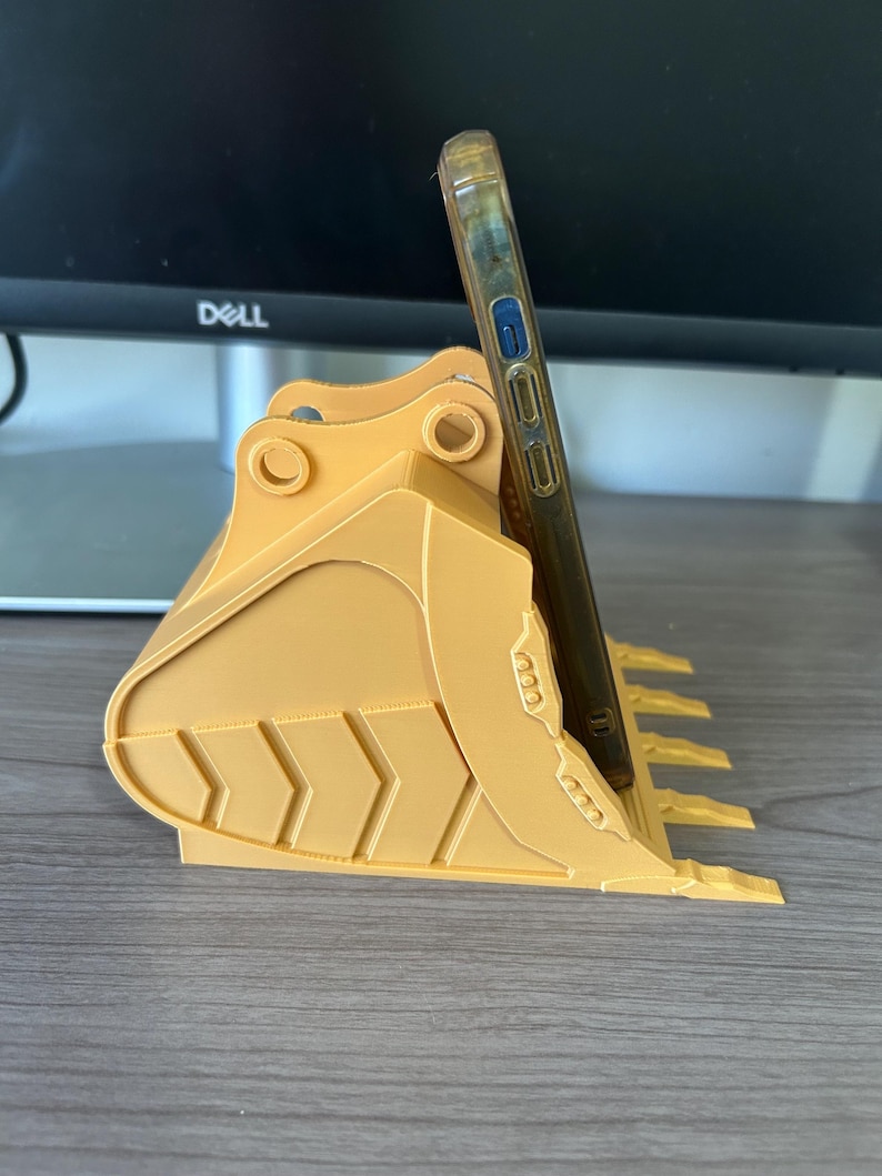 Puede incluir: Un soporte para tel&eacute;fono amarillo impreso en 3D con forma de cuchara de excavadora. Tiene una ranura para sujetar un tel&eacute;fono en posici&oacute;n vertical. El tel&eacute;fono est&aacute; en una funda transparente. El soporte est&aacute; sobre una superficie de madera.