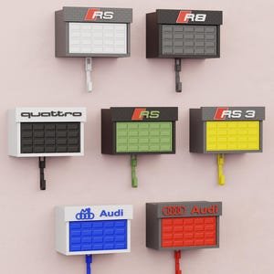 May include: A collection of wall-mounted key holders in various colors, each featuring a different Audi logo or model designation, such as "RS" and "quattro." The key holders have a rectangular design with a hook below.
