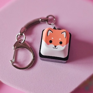 Könnte beinhalten: Ein quadratischer Schlüsselanhänger mit einem Shiba Inu-Hunde-Gesicht in Orange, Weiß und Schwarz. Der Anhänger ist an einem silberfarbenen Schlüsselring und einem geflochtenen grauen Riemen befestigt. Der Anhänger wird auf einer rosa sechseckigen Oberfläche vor einem rosa Hintergrund präsentiert.