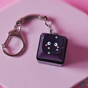 Può includere: Un portachiavi viola e nero con chiusura argentata. Il portachiavi presenta un ciondolo quadrato con un volto di cartone animato e dettagli a stella. Il ciondolo è attaccato a un anello di metallo argentato e a una chiusura. Lo sfondo è rosa con decorazioni a forma di stella.