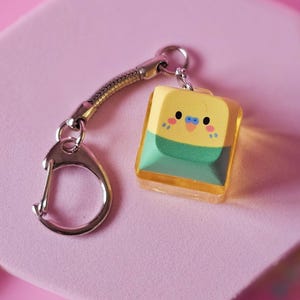 May include: A yellow and green bird-shaped keyring charm with a silver clasp. The charm is a small, rectangular shape with a cartoon bird design. The background is a pink hexagonal shape with colourful star-shaped decorations.