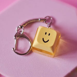 Peut inclure: Un porte-clés en forme de touche de clavier jaune avec un visage souriant. Le porte-clés a un fermoir argenté et une chaîne argentée enroulée. La clé est sur une plateforme hexagonale rose, avec un fond rose et des décorations en forme d'étoiles.