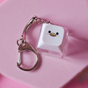 以下が含まれることがあります： 白いキーボードのキーに、漫画のアヒルの顔のデザインが付いたシルバーのキーホルダー。 キーホルダーはピンク色の六角形の表面にあり、ピンク色の背景と星型の装飾が施されています。