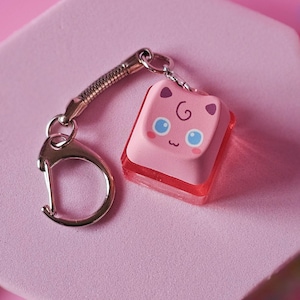 May include: A pink keychain featuring a cartoon character design. The character has blue eyes and a swirl on its forehead. The keychain is attached to a silver-colored clasp and a pink, hexagonal base.