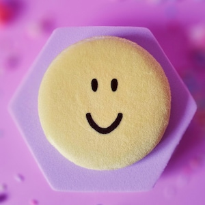May include: A yellow smiley face on a hexagonal lavender base, set against a soft purple background. The smiley has black eyes and a curved mouth. Scattered confetti adds a playful touch to the composition.