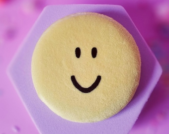 May include: A yellow smiley face on a hexagonal lavender base, set against a soft purple background. The smiley has black eyes and a curved mouth. Scattered confetti adds a playful touch to the composition.