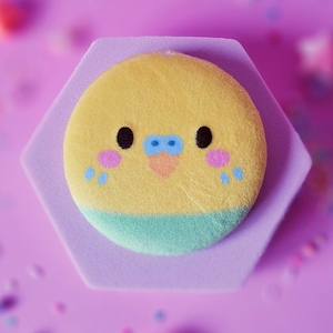 Op de afbeelding: Een ronde, gele pluche met een vogelgezichtontwerp, met zwarte ogen, roze wangen en een blauwe en oranje snavel. De pluche zit op een paarse zeshoekige basis, omringd door kleurrijke confetti op een paarse achtergrond.