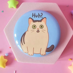 Könnte beinhalten: Ein runder, hellblauer Button mit einer Cartoon-Katzen-Illustration. Die Katze ist beige und braun mit großen gelben Augen und dem Wort "Huh?" darüber. Der Button befindet sich auf einer rosa sechseckigen Basis, umgeben von bunten stern- und kugelförmigen Dekorationen.
