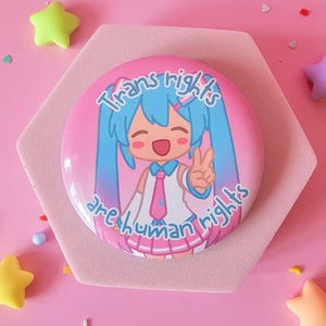 Puede incluir: Un botón redondo rosa con el texto "Trans rights are human rights" y un personaje de estilo anime con cabello azul. El botón está sobre un soporte hexagonal rosa, rodeado de estrellas y otras decoraciones coloridas.